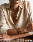 Person wearing a beige shirt with a necklace and bracelets, sitting on a woven chair.