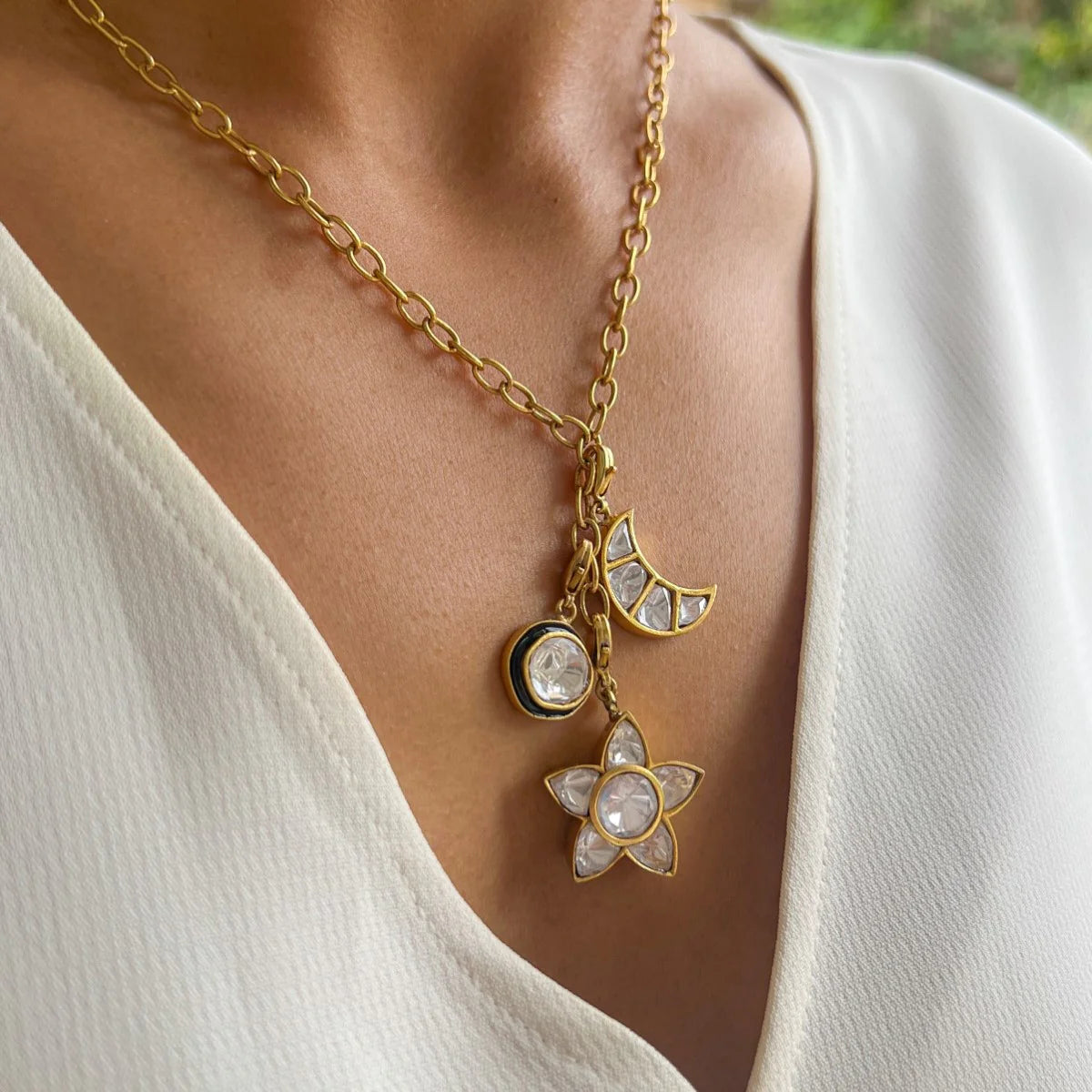 A gold-colored necklace with three charm pendants in the shapes of a moon, a star, and a circular disc, worn by a person in a white V-neck top.