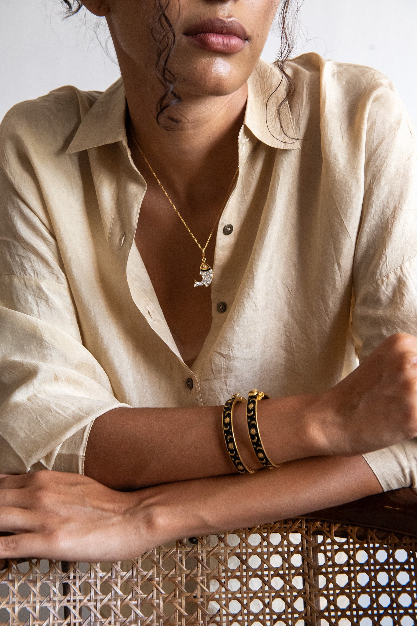 Person wearing a beige shirt with a necklace and bracelets, sitting on a woven chair.