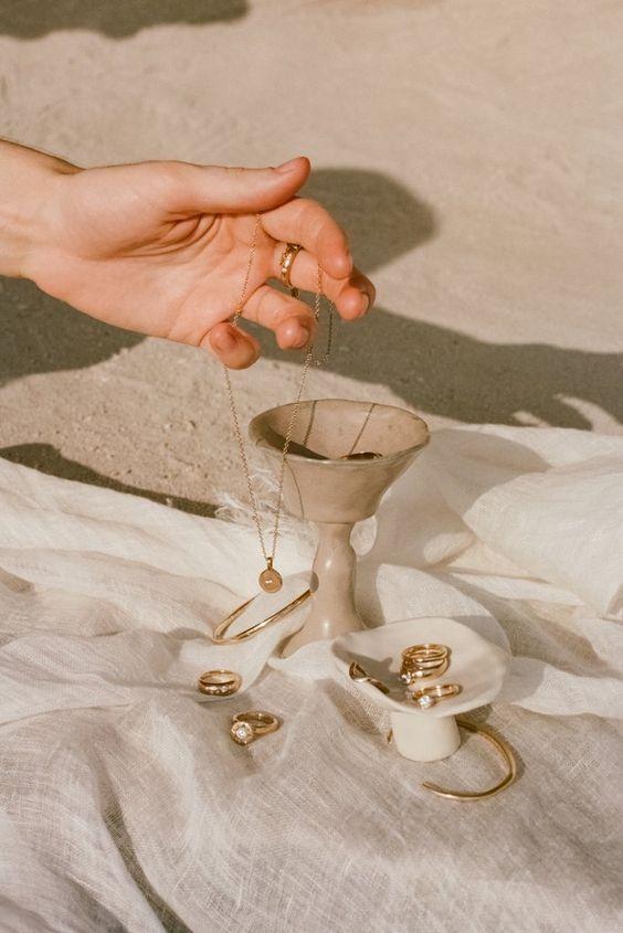 Hand holding a necklace over a small white stand with rings on a light fabric background